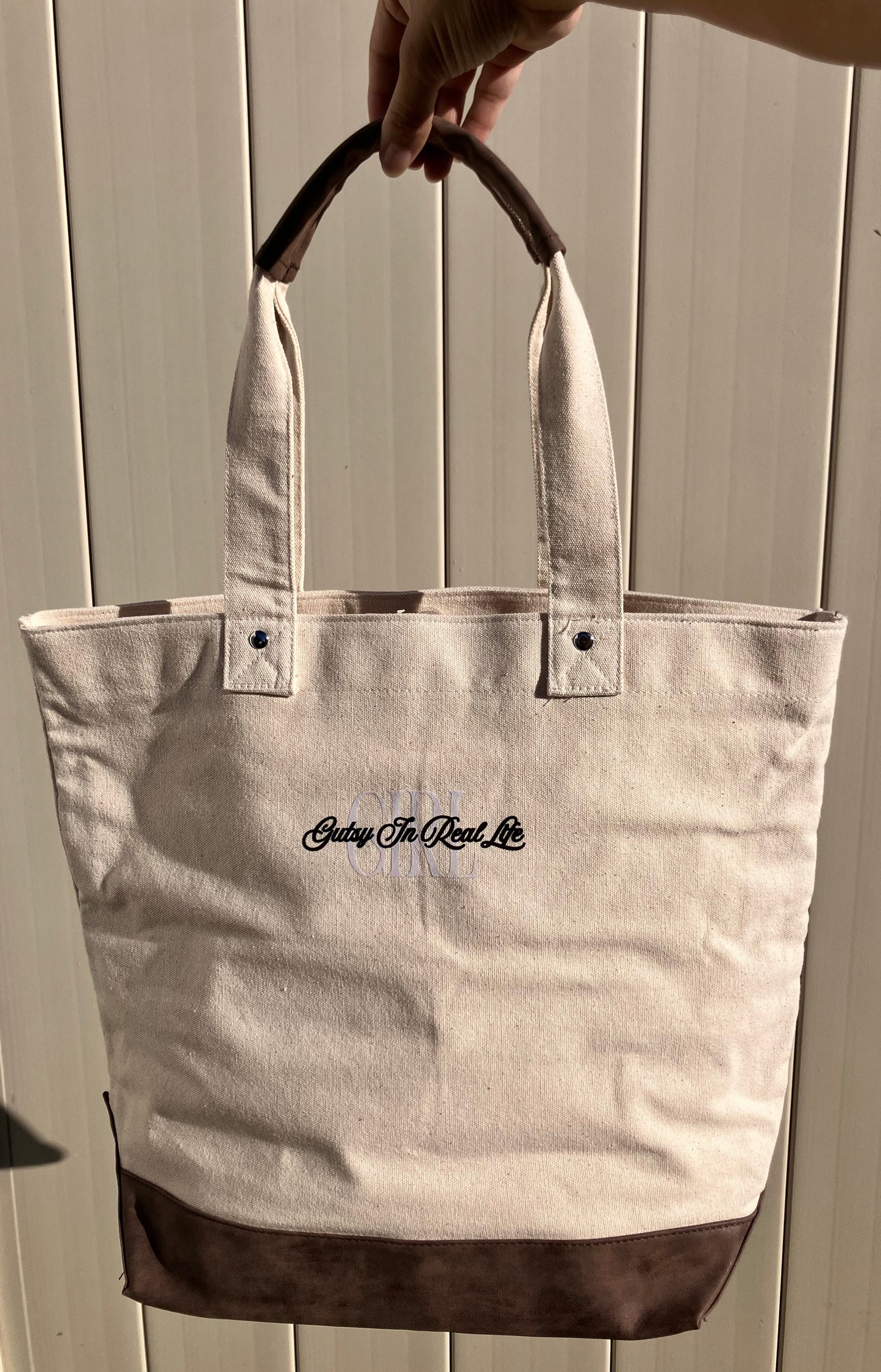 Beige tote bag with brown accents held by a hand against a white background