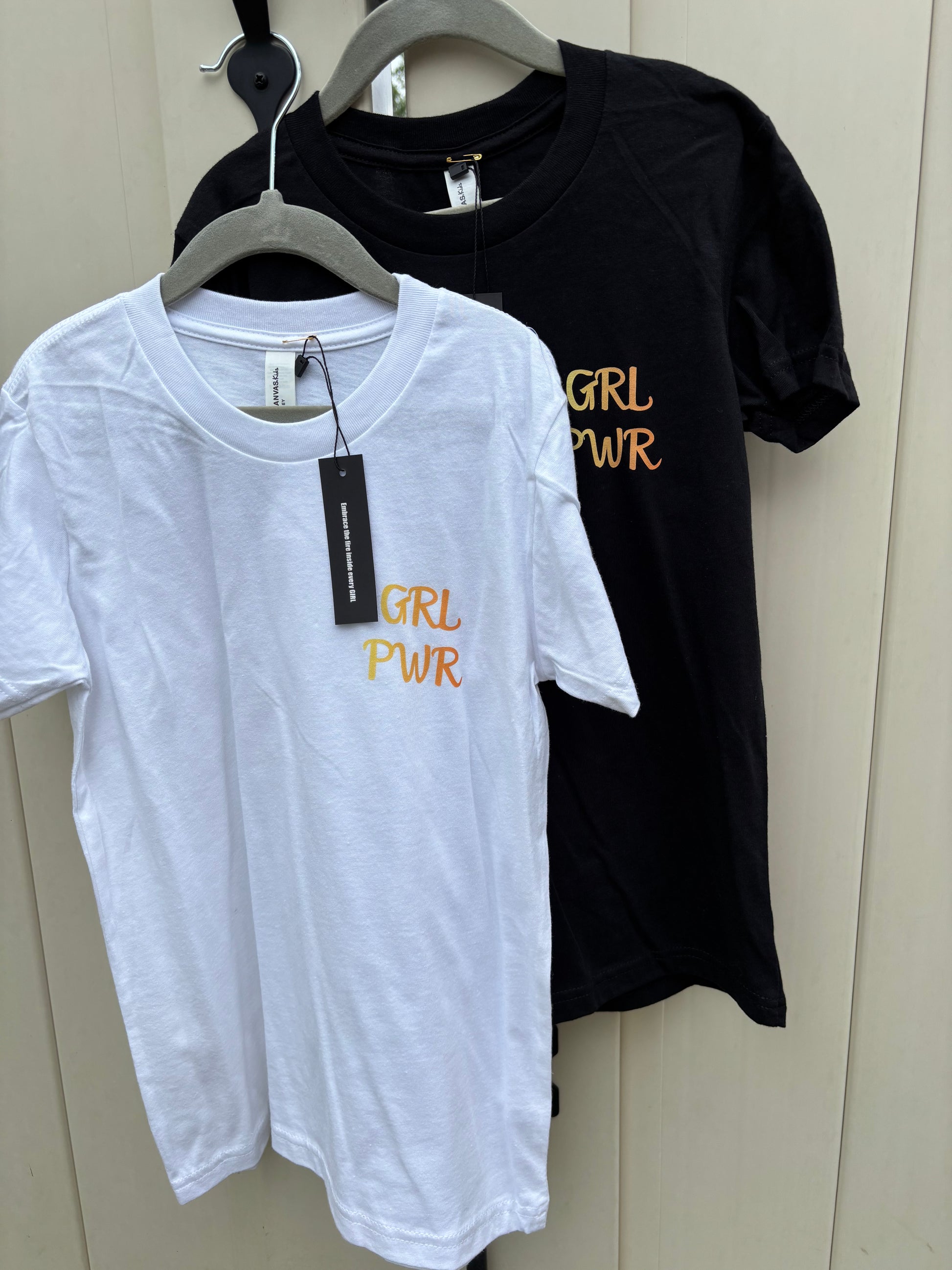 Black and white t-shirts with 'GRL PWR' text on hangers against a neutral background