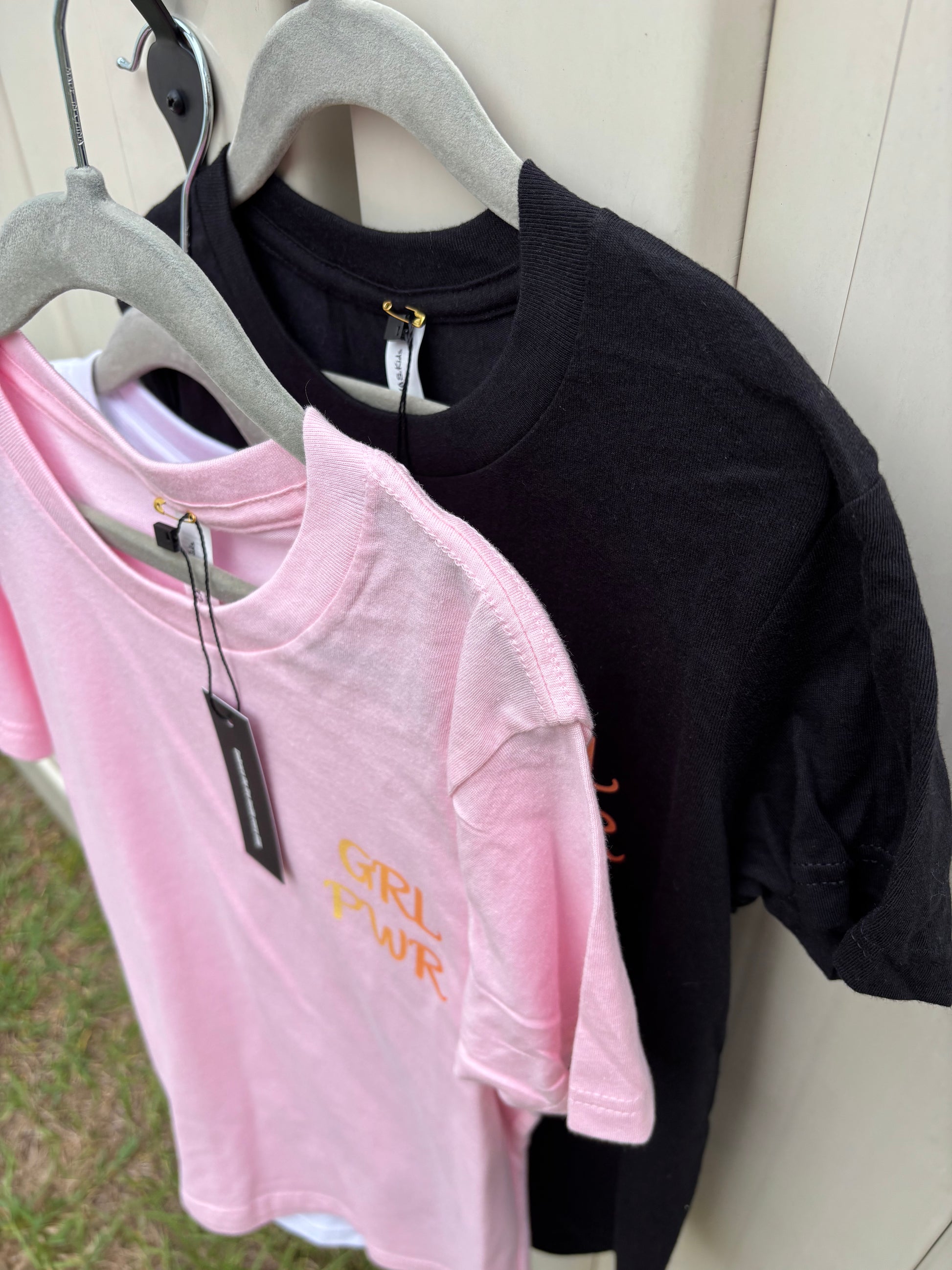 Black and pink t-shirts on hangers with text on a wooden surface.