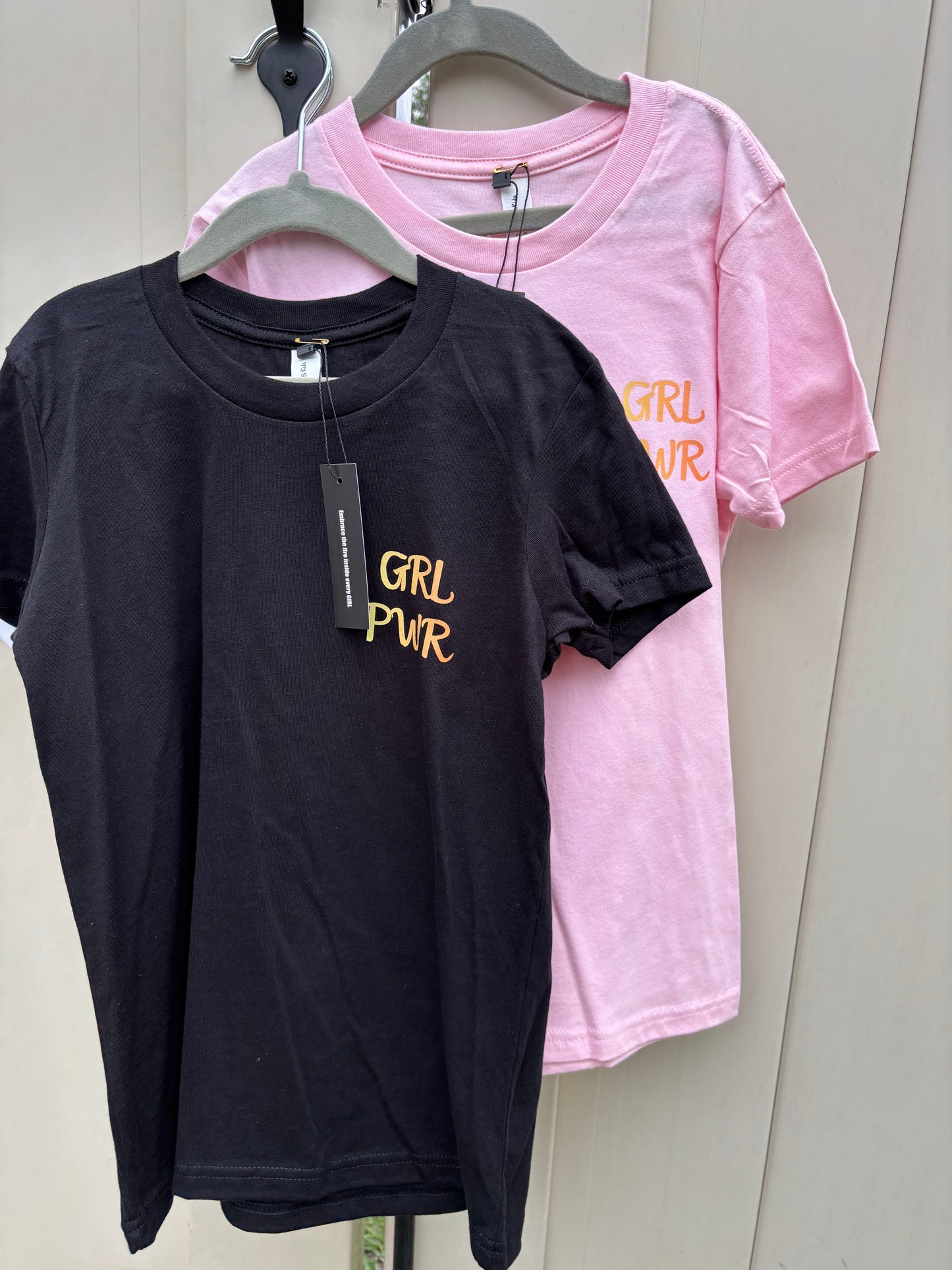 Black t-shirt with 'Girl Power' text on a hanger against a pink shirt background.
