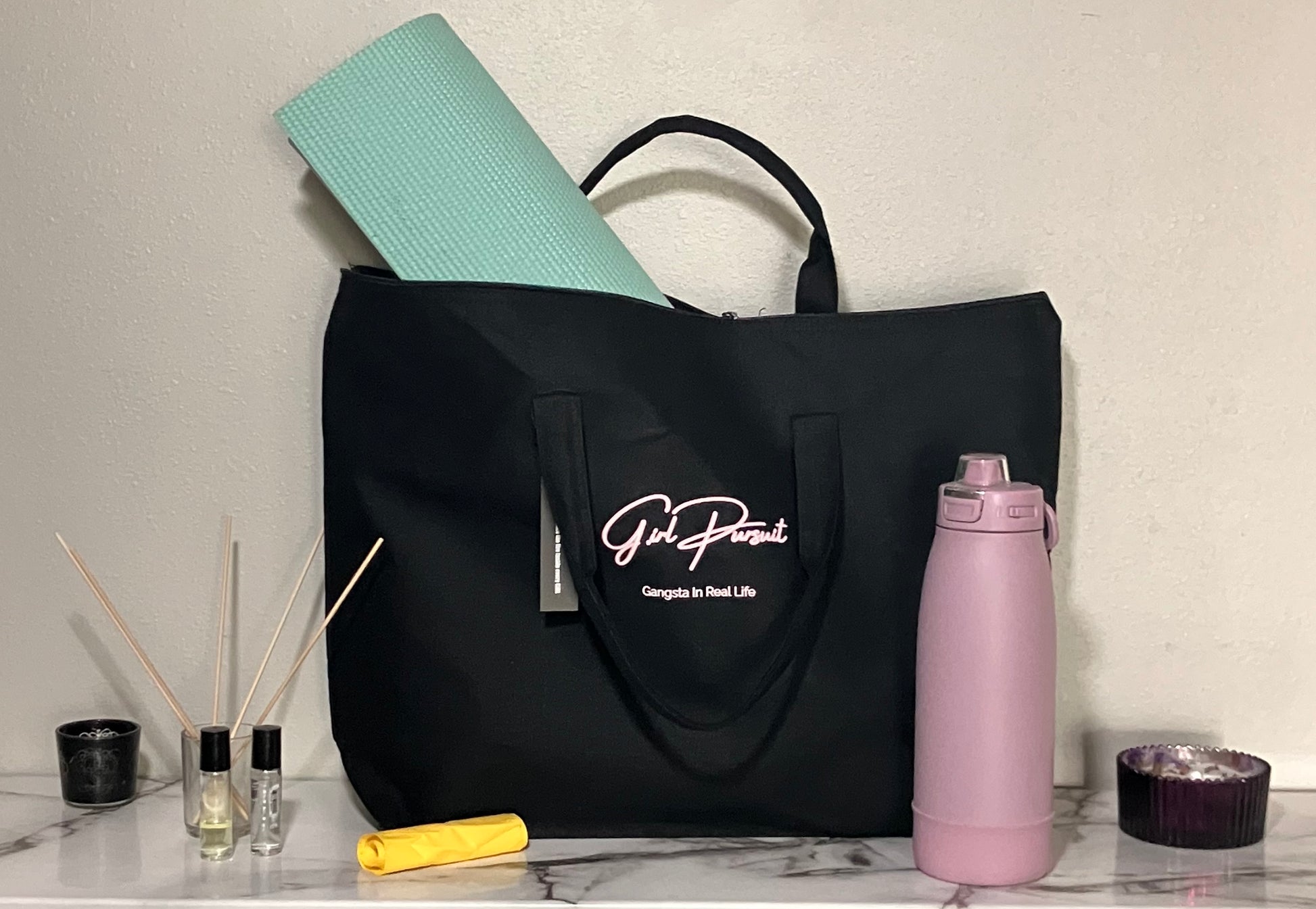 Black tote bag with a brand logo, yoga mat, pink water bottle, and small items on a marble surface.