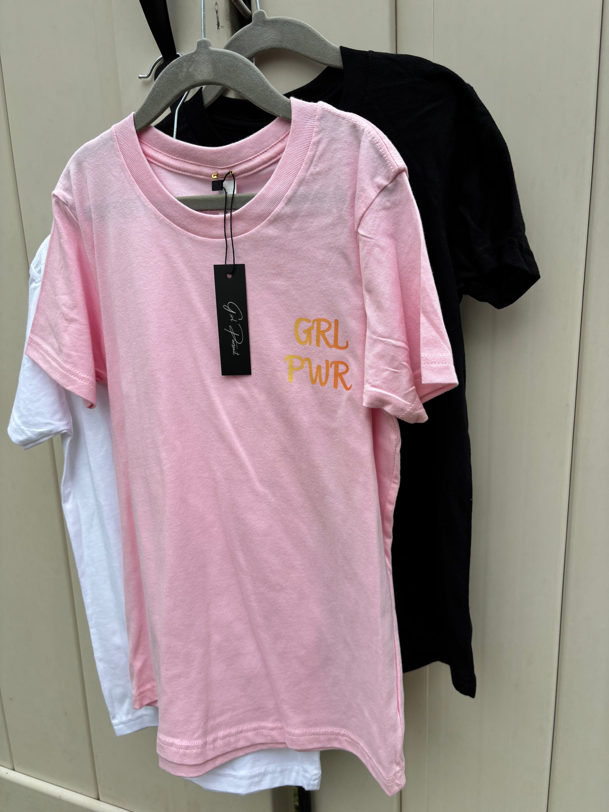 Pink t-shirt with 'GRL PWR' text on a hanger against a neutral background