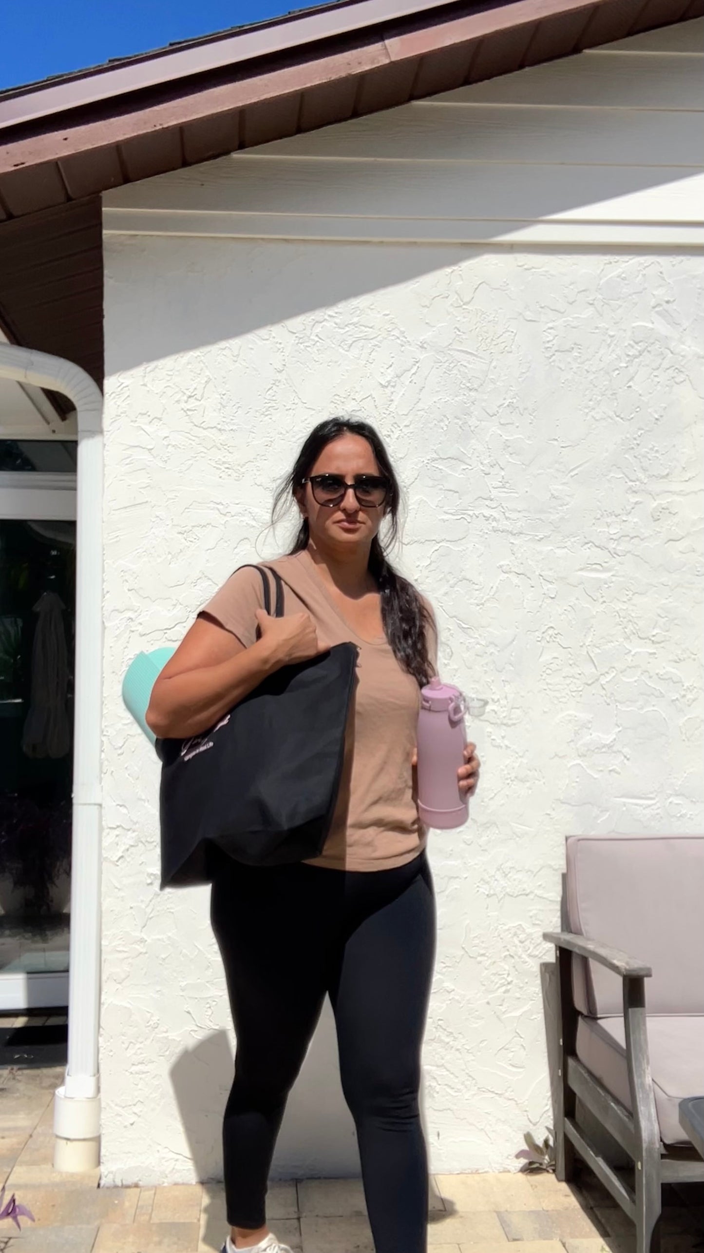 Woman holding a black bag and pink water bottle in front of a white building.