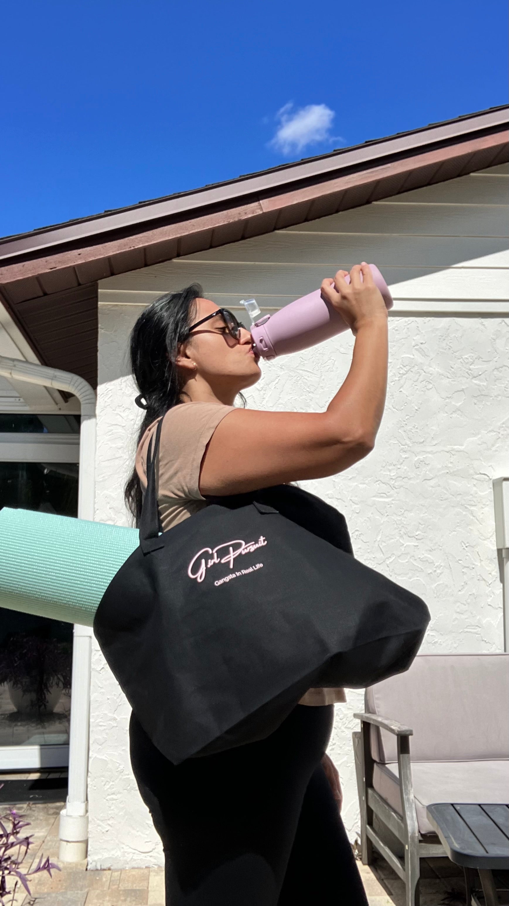 Person holding a black bag with a logo, standing outdoors on a sunny day with a Yoga Mat in the bag.
